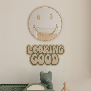 May include: A round smiley face mirror and a wall sign with the text LOOKING GOOD in green and beige. Below, a sage green toy car, a small grey bear figurine, a wooden mannequin, and a green plastic storage crate sit on a beige cabinet.