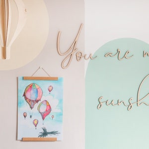 May include: A light-coloured wooden hot air balloon wall decoration, a vertical poster with a watercolour painting of colourful hot air balloons, and wooden script lettering that reads "You are my sunshine" mounted on a wall.