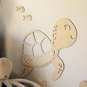Puede incluir: Decoración de pared de madera con una tortuga sonriente, peces, olas y conchas marinas. La tortuga tiene un diseño detallado de caparazón. La decoración es de madera clara, creando un tema náutico para una habitación infantil.