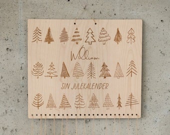 May include: A rectangular light-colored wooden board featuring engraved illustrations of various pine trees, the name William, and the text SIN JULEKALENDER. Multiple brown paper house-shaped boxes hang from the board by thin twine strings.