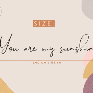 May include: A wall sign featuring the black cursive text You are my sunshine. The sign measures 33 cm in height and 140 cm in width. The background is a light beige colour with abstract shapes in pink, yellow, and purple. A logo with a flamingo is in the top left corner.