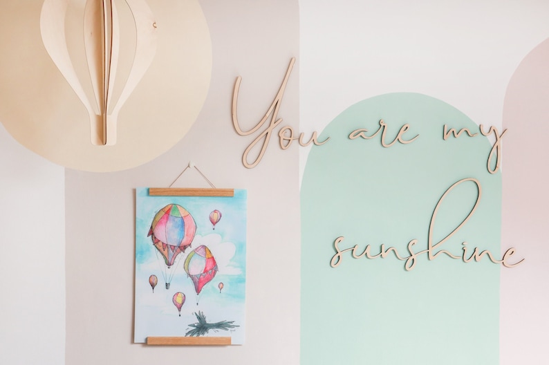 May include: A light-coloured wooden hot air balloon wall decoration, a vertical poster with a watercolour painting of colourful hot air balloons, and wooden script lettering that reads "You are my sunshine" mounted on a wall.