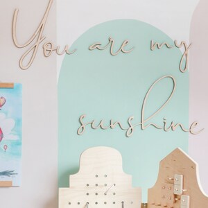 May include: A light-coloured wooden wall sign featuring the cursive text "You are my sunshine" in two lines. Below the sign are two light-coloured wooden pegboards with various small wooden shapes and grey strings attached to the perforated surfaces.