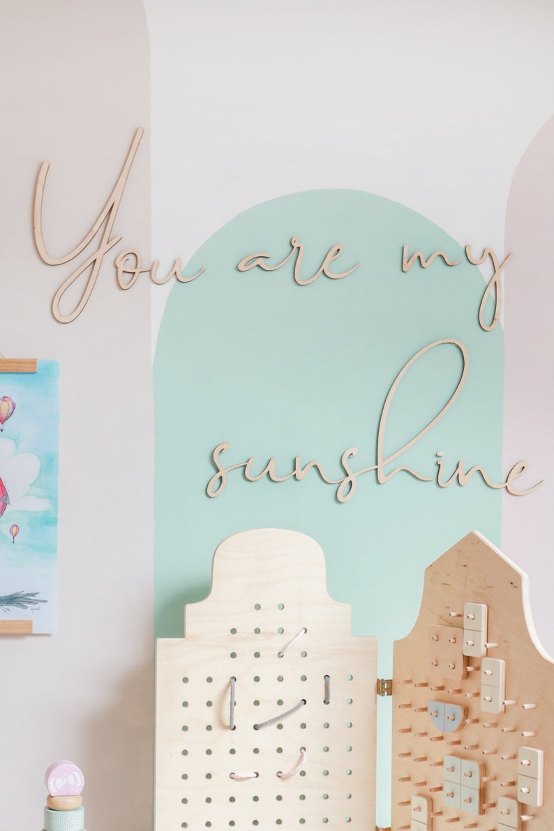 May include: A light-coloured wooden wall sign featuring the cursive text "You are my sunshine" in two lines. Below the sign are two light-coloured wooden pegboards with various small wooden shapes and grey strings attached to the perforated surfaces.
