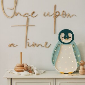 May include: A wooden wall sign with the text Once upon a time in cursive script, accompanied by a penguin-shaped LED lamp with a dark teal head, light teal wings, and a white belly with small glowing lights, plus wooden toys on a white cabinet.