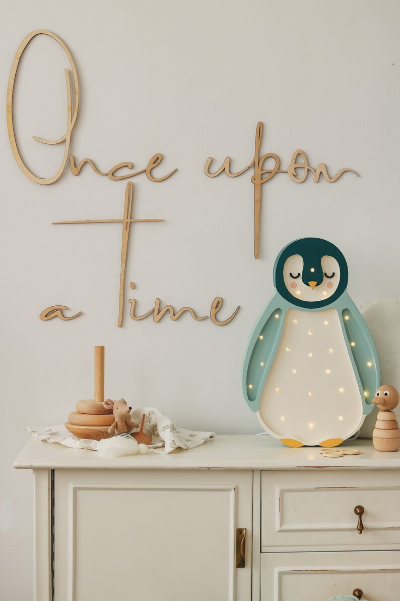 May include: A wooden wall sign with the text Once upon a time in cursive script, accompanied by a penguin-shaped LED lamp with a dark teal head, light teal wings, and a white belly with small glowing lights, plus wooden toys on a white cabinet.