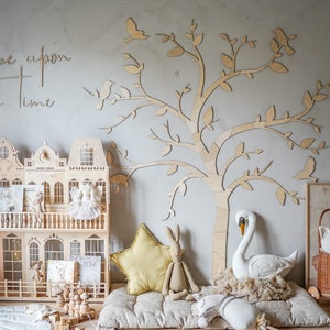 May include: A wooden dollhouse with architectural details, a large laser-cut wooden tree wall decal, and the text "Once upon a time" on the wall. The scene includes a yellow star pillow, a plush rabbit, a white swan, and a wicker basket with a cat doll.