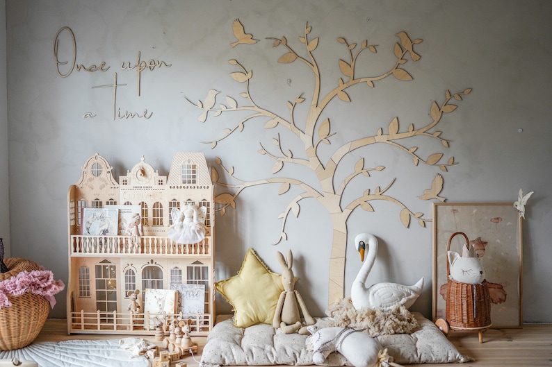 May include: A wooden dollhouse with architectural details, a large laser-cut wooden tree wall decal, and the text "Once upon a time" on the wall. The scene includes a yellow star pillow, a plush rabbit, a white swan, and a wicker basket with a cat doll.