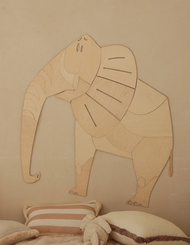 Op de afbeelding: Een houten olifantenwanddecoratie in een lichte kleur. De olifant heeft een segmentontwerp, met een lange slurf en grote oren. Het is gemonteerd op een neutrale muur boven kussens.