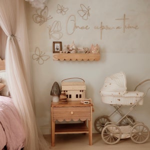 May include: A nursery room featuring a wooden nightstand with a scalloped edge, a toy house, a wooden stacking toy, and a mushroom-shaped lamp. A vintage-style doll carriage sits nearby. The wall displays wire butterfly art and the text Once upon a time.