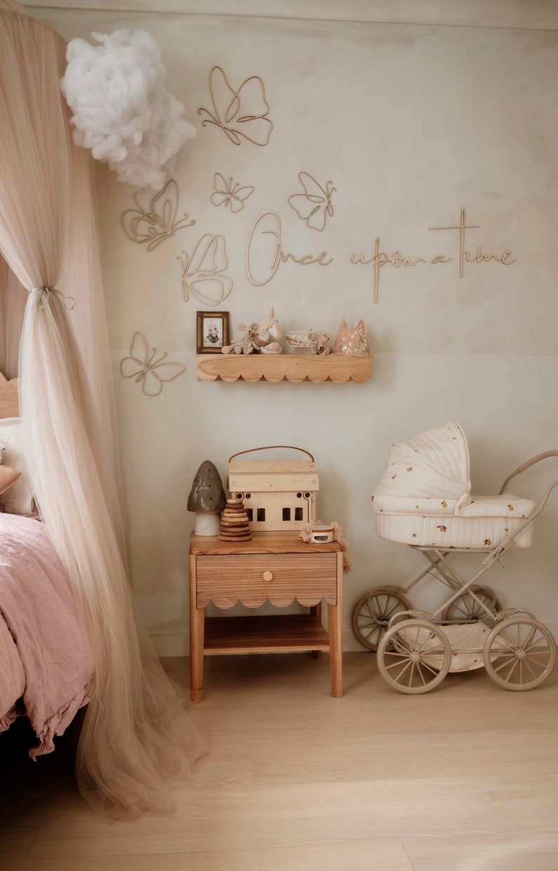 May include: A nursery room featuring a wooden nightstand with a scalloped edge, a toy house, a wooden stacking toy, and a mushroom-shaped lamp. A vintage-style doll carriage sits nearby. The wall displays wire butterfly art and the text Once upon a time.