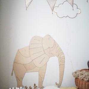 Op de afbeelding: Een houten olifantenwanddecoratie in een kinderkamer, aangevuld met een slinger van wimpels en een wolkvormige lamp. Een kegelspel en houten speelgoed liggen op de vloer.
