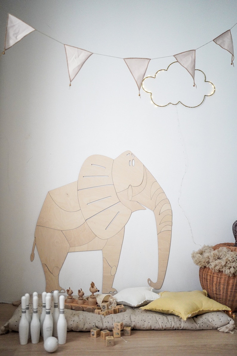 Op de afbeelding: Een houten olifantenwanddecoratie in een kinderkamer, aangevuld met een slinger van wimpels en een wolkvormige lamp. Een kegelspel en houten speelgoed liggen op de vloer.