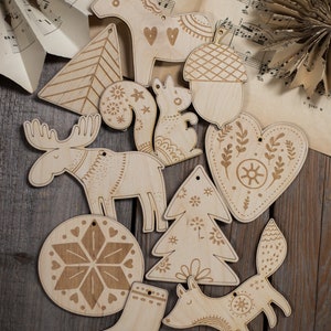 May include: A collection of light-colored wooden ornaments featuring laser-engraved designs including a moose, a fox, a squirrel, an acorn, a heart, a stocking, a snowflake, and various tree shapes, each with a small hole for hanging.