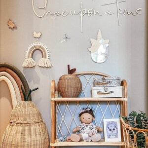May include: A rattan shelf unit holds a doll, two small suitcases, and a framed print. Beside it sits a large pear-shaped woven basket. The wall features a wooden script sign reading Once upon a time, a rainbow wall hanging, and a unicorn mirror.