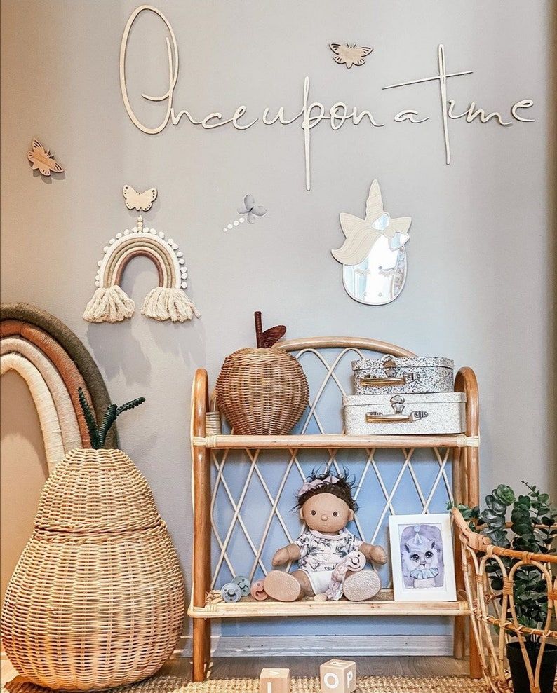 May include: A rattan shelf unit holds a doll, two small suitcases, and a framed print. Beside it sits a large pear-shaped woven basket. The wall features a wooden script sign reading Once upon a time, a rainbow wall hanging, and a unicorn mirror.