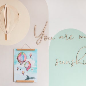 May include: A wooden hot air balloon wall decoration, a hanging poster featuring a watercolour illustration of colourful hot air balloons, and wooden script lettering that reads You are my sunshine, all displayed on a wall with painted arches.