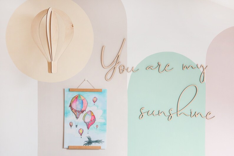 May include: A wooden hot air balloon wall decoration, a hanging poster featuring a watercolour illustration of colourful hot air balloons, and wooden script lettering that reads You are my sunshine, all displayed on a wall with painted arches.