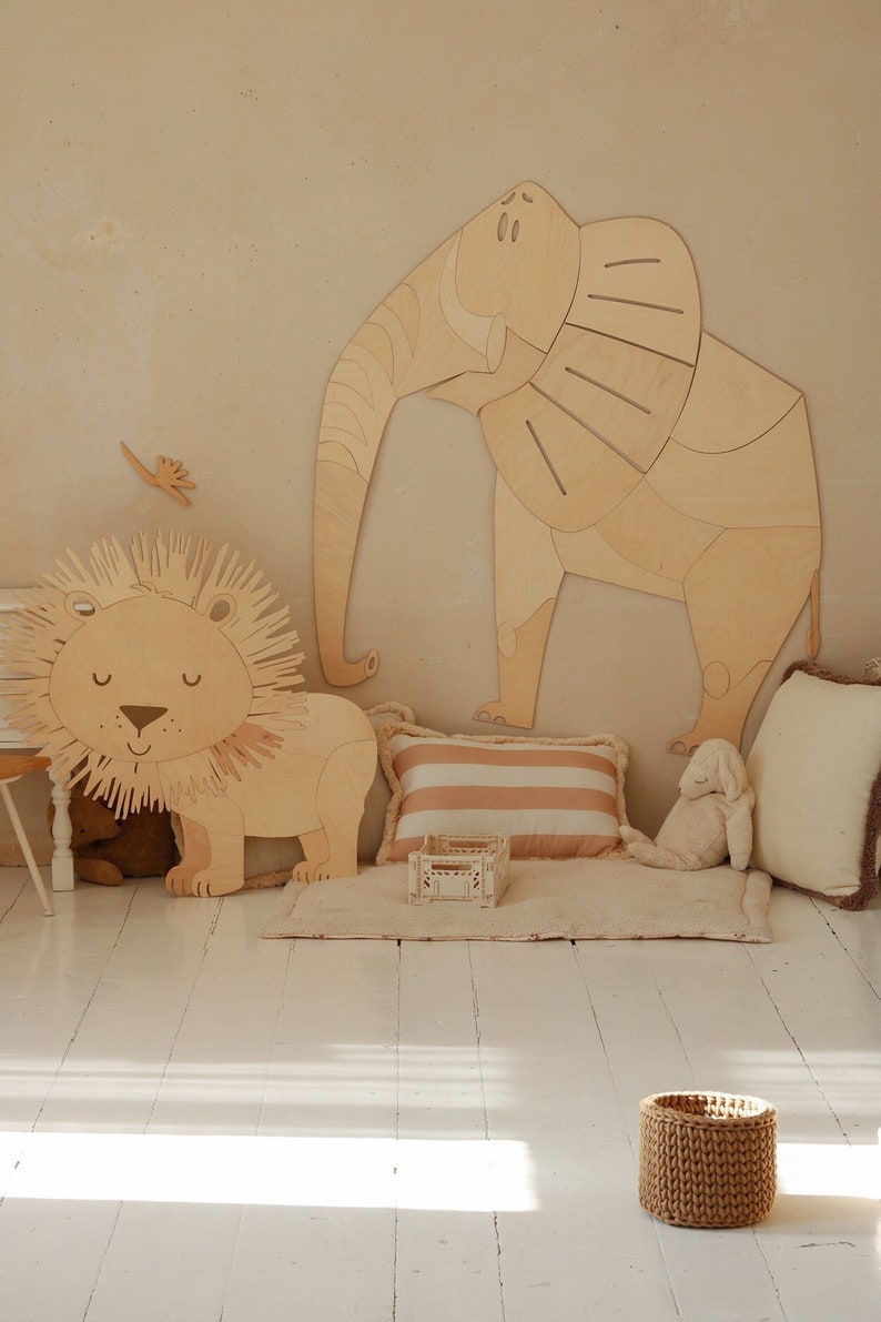 Op de afbeelding: Houten wanddecoratie van een leeuw en een olifant in een kinderkamer. De leeuw heeft een gedetailleerde manen en de olifant heeft grote oren. Een gestreept kussen, een kleine houten kist en een gevlochten mand zijn ook aanwezig.