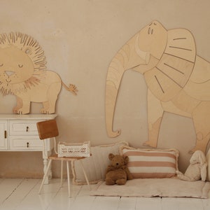 Op de afbeelding: Houten wanddecoratie van een leeuw en een olifant. De leeuw heeft een gedetailleerde manen en de olifant grote oren. De kamer heeft ook een witte kast, een kleine stoel, kussens en een teddybeer.