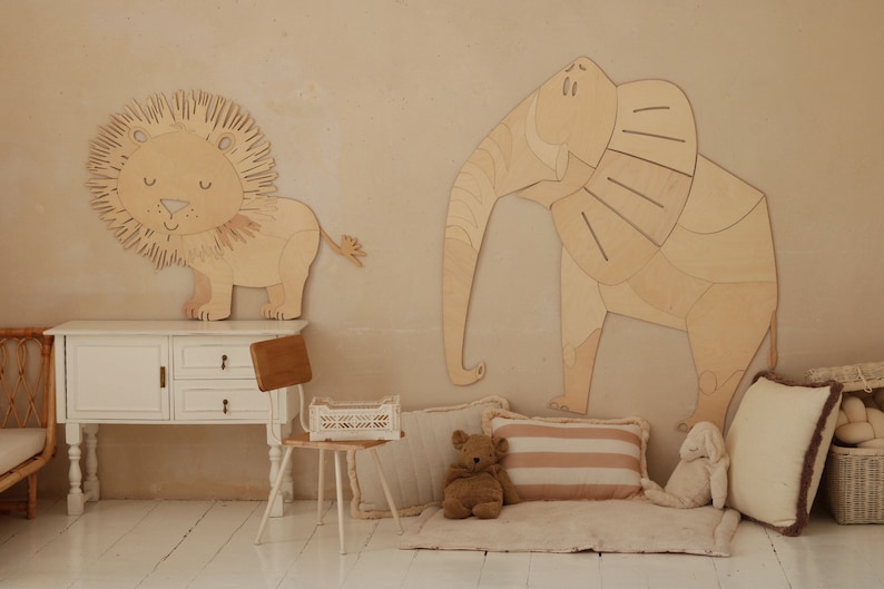 Op de afbeelding: Houten wanddecoratie van een leeuw en een olifant. De leeuw heeft een gedetailleerde manen en de olifant grote oren. De kamer heeft ook een witte kast, een kleine stoel, kussens en een teddybeer.