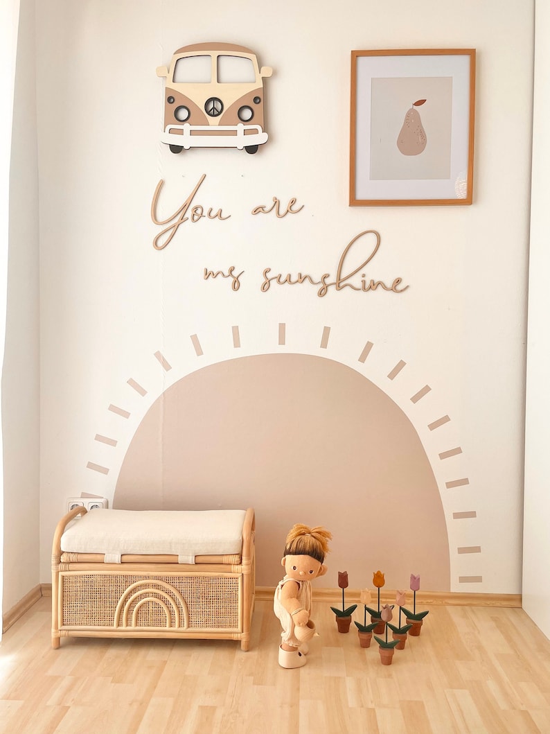 May include: A room corner featuring a beige and white wall decal of a sun, a wooden camper van wall hanging, a framed pear print, and the text "You are my sunshine". A rattan storage bench with a white cushion and a doll with toy flowers are present.