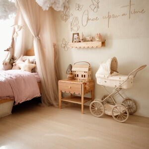 May include: A bedroom featuring a bed with a beige canopy, a wooden nightstand with a scalloped edge, and a vintage-style doll carriage. The wall displays butterfly wall art and the text Once upon a time in a light wood finish.