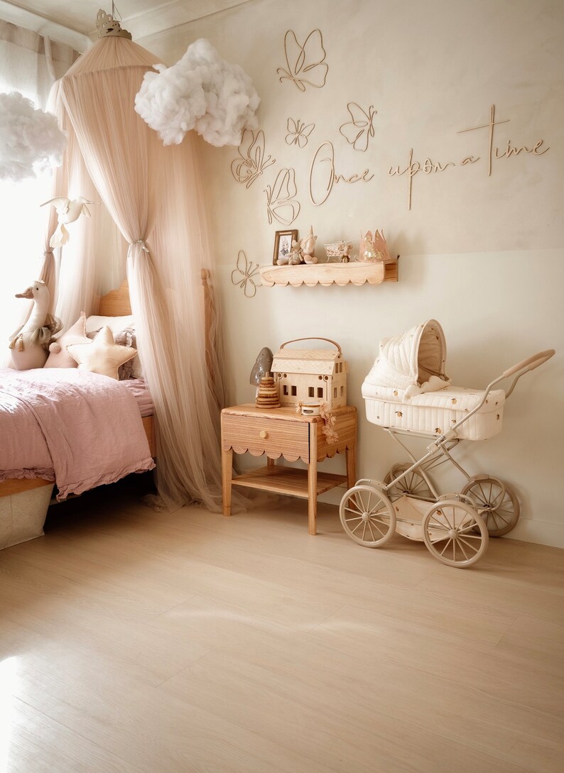 May include: A bedroom featuring a bed with a beige canopy, a wooden nightstand with a scalloped edge, and a vintage-style doll carriage. The wall displays butterfly wall art and the text Once upon a time in a light wood finish.