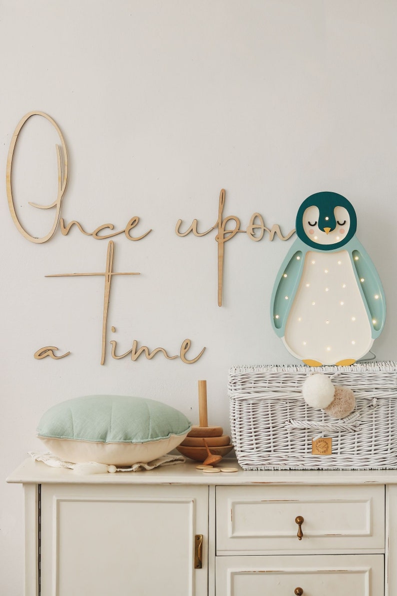 May include: A wooden wall sign with the cursive text Once upon a time, a penguin-shaped LED wall lamp with teal and white accents, a round mint green pillow, a wooden stacking toy, and a white woven basket with two pom-poms on top.