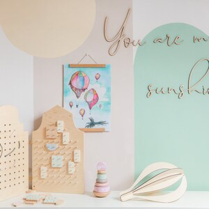 May include: Two light-coloured wooden pegboards with holes, a watercolour print of hot air balloons, a wooden stacking toy with pastel rings, and a wooden hot air balloon sculpture. The wall features the wooden script text You are my sunshine.
