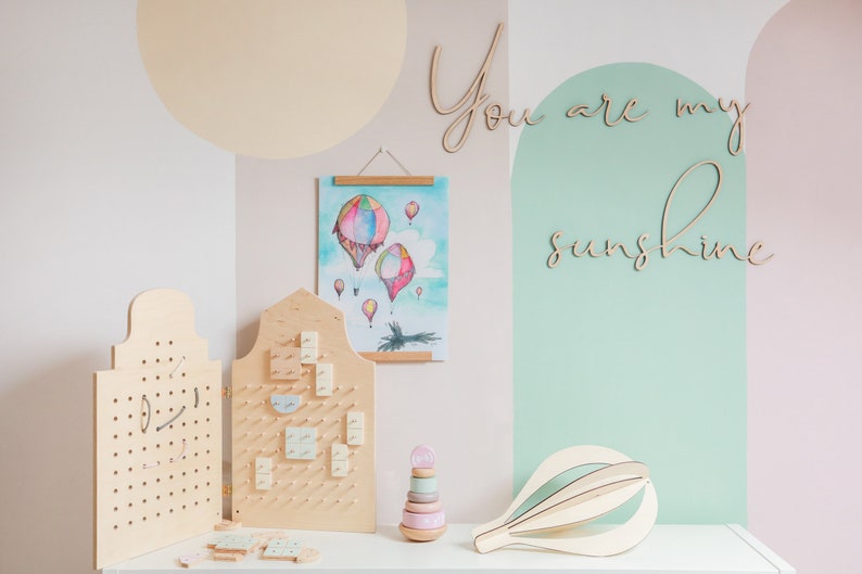 May include: Two light-coloured wooden pegboards with holes, a watercolour print of hot air balloons, a wooden stacking toy with pastel rings, and a wooden hot air balloon sculpture. The wall features the wooden script text You are my sunshine.