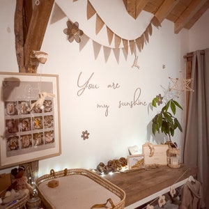 May include: A nursery room featuring a wooden changing table with a woven basket tray, a framed poster, and wall decor including a brown fabric bunting, wooden flower cutouts, and the script text "You are my sunshine" in wooden letters.