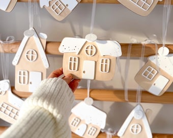 May include: A collection of wooden house-shaped ornaments featuring white roofs with speckled details, white doors, and white window frames. Each ornament is suspended by a white ribbon from a wooden rack, with a small numbered tag on each piece.