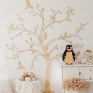 May include: A large, flat, light-colored wooden tree wall decal with branches and leaves, featuring bird silhouettes. A black and tan wooden penguin figure sits on a white cabinet next to a beige drawstring bag with text.