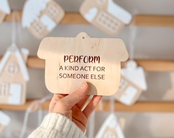 May include: A light-coloured wooden ornament shaped like a house with a small hole at the top for hanging. The text "PERFORM A KIND ACT FOR SOMEONE ELSE" is engraved in dark brown letters on the front surface of the smooth wooden house shape.