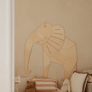 Op de afbeelding: Een groot houten olifantenkunstwerk voor aan de muur is bevestigd aan een beige muur. Daaronder ondersteunt een pluche tapijt een teddybeer, een gestreept kussen en een wit kussen met een bruine rand.