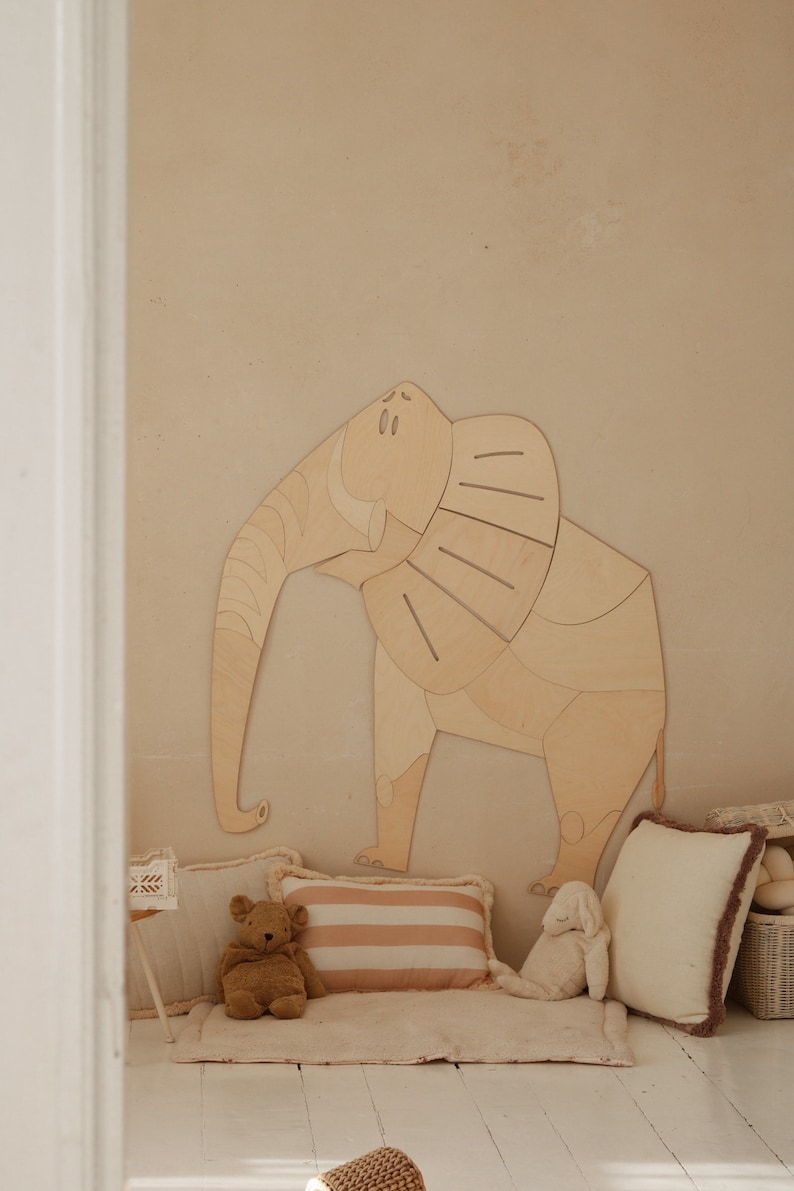 Op de afbeelding: Een groot houten olifantenkunstwerk voor aan de muur is bevestigd aan een beige muur. Daaronder ondersteunt een pluche tapijt een teddybeer, een gestreept kussen en een wit kussen met een bruine rand.