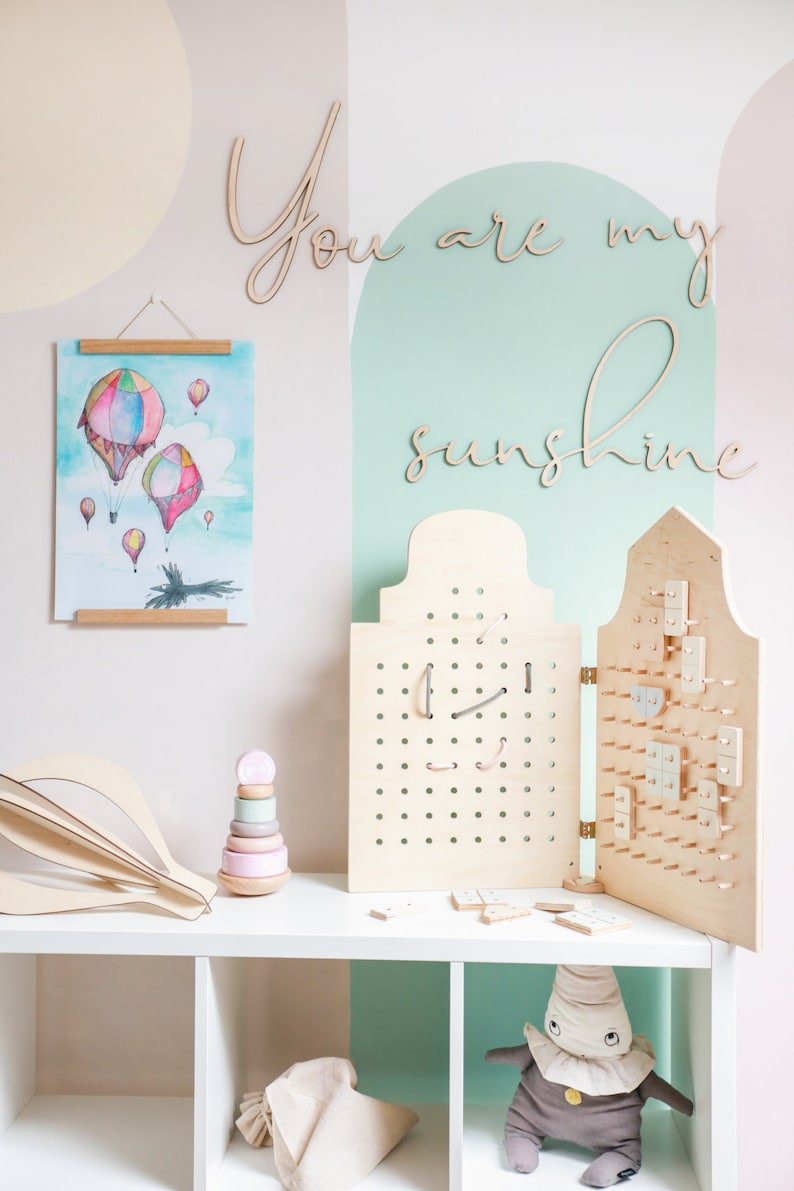 May include: A wooden pegboard activity set with two hinged panels, featuring various small wooden shapes attached to the board. Beside it sits a pastel-coloured stacking ring toy, a watercolour print of hot air balloons, and the wooden text You are my sunshine.