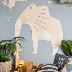 Op de afbeelding: Houten olifant muurkunst met een toekan en boom uitsparingen op een lichtblauwe muur. Een hond rust op een gestreept kussen bij een girafvormige lamp. Een kussen leest "Stay Wild Child".