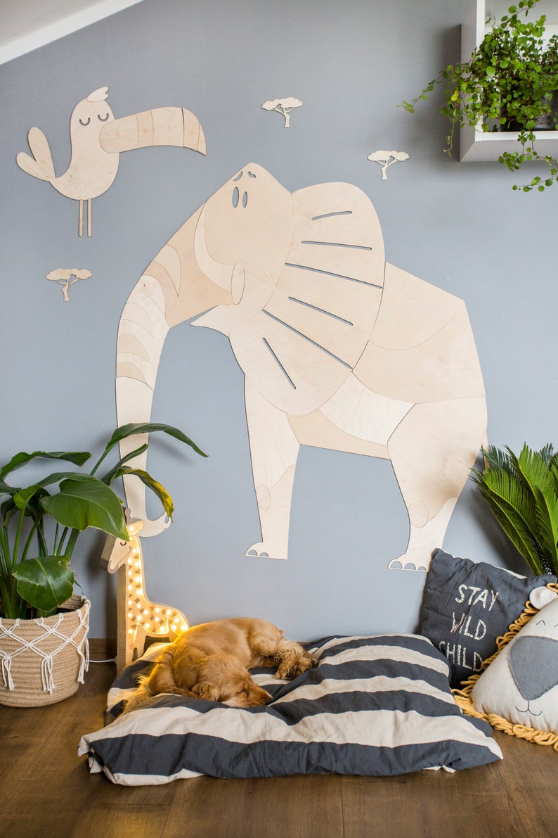 Op de afbeelding: Houten olifant muurkunst met een toekan en boom uitsparingen op een lichtblauwe muur. Een hond rust op een gestreept kussen bij een girafvormige lamp. Een kussen leest "Stay Wild Child".
