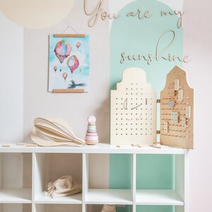May include: A white shelving unit displays a wooden hot air balloon sculpture, a pastel stacking toy, and a folding wooden pegboard with various small wooden shapes. A watercolour print of hot air balloons hangs above, with the wooden text You are my sunshine.