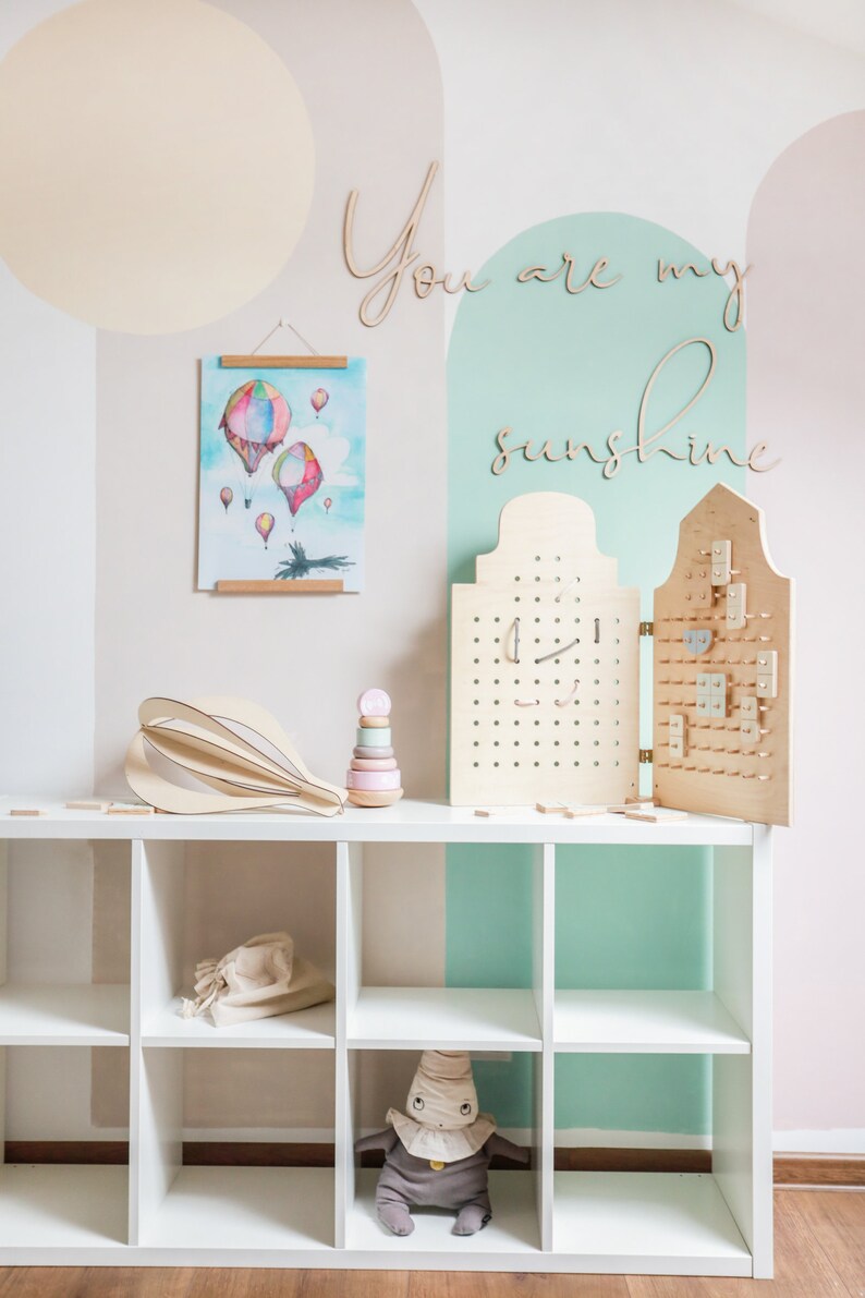 May include: A white shelving unit displays a wooden hot air balloon sculpture, a pastel stacking toy, and a folding wooden pegboard with various small wooden shapes. A watercolour print of hot air balloons hangs above, with the wooden text You are my sunshine.