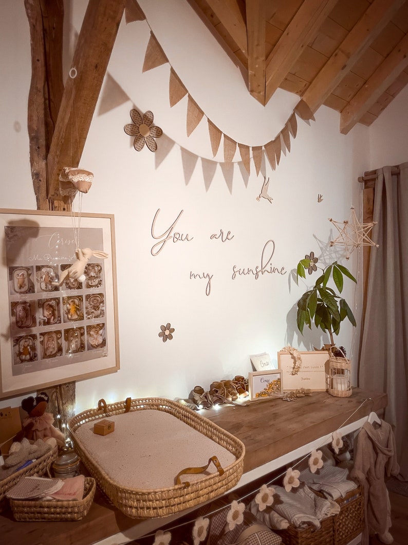 May include: A nursery room featuring a wooden changing table with a woven basket tray, a framed poster, and wall decor including a brown fabric bunting, wooden flower cutouts, and the script text "You are my sunshine" in wooden letters.
