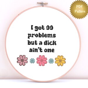 May include: Cross stitch pattern featuring the text "I got 99 problems but a dick ain't one" with a row of pink, blue, and yellow flowers at the bottom.