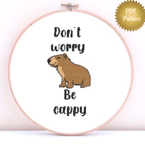May include: Cross stitch pattern featuring a brown capybara with the text "Don't worry be cappy" in black.