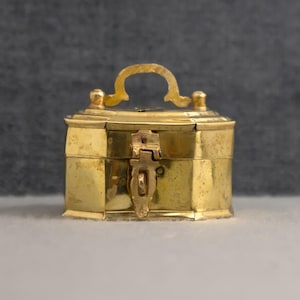 以下が含まれることがあります： 楕円形の小さな真鍮製の箱で、ヒンジ付きの蓋が付いています。箱の上部には装飾的なアーチ型のハンドルがあり、前面には中央にラッチ機構が備わっています。金属の表面は温かみのある金色で、わずかな使用感が見られます。