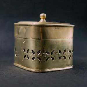 Könnte beinhalten: Eine quadratische, messingfarbene Box mit Deckel und kleinem Knopf oben. Die Seiten der Box weisen ein dekoratives Muster aus ausgeschnittenen Blumenformen auf. Die Box wird wahrscheinlich zur Aufbewahrung oder Präsentation verwendet.