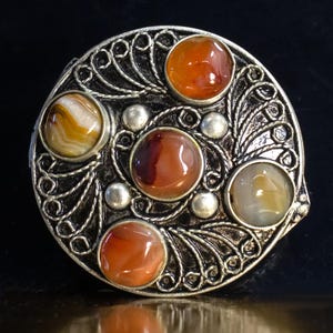 Puede incluir: Broche redondo plateado con un diseño ornamentado. Presenta cinco piedras preciosas pulidas en tonos naranja, marrón y crema, engastadas en un marco metálico detallado y arremolinado. Pequeñas cuentas plateadas acentúan el diseño.