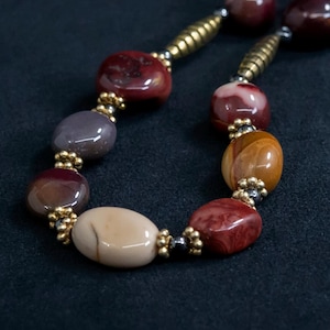 Op de afbeelding: Een close-up van een ketting met grote, gepolijste kralen in tinten rood, bruin en beige. De kralen worden gescheiden door kleine goudkleurige afstandhouders en verbonden door een goudkleurige ketting. De ketting wordt tegen een donkere achtergrond getoond.