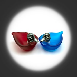 Op de afbeelding: Twee glazen decoratieve objecten in de vorm van een eend. Eén is dieprood van kleur en de andere is helderblauw. Elk heeft een zilverkleurige eendenkop als stopper. De objecten staan op een witte ondergrond.
